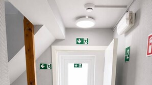 A narrow hallway with a wooden staircase, a door with an emergency exit sign, and a wall-mounted fire extinguisher. - Hotel Albergo (Brussels)