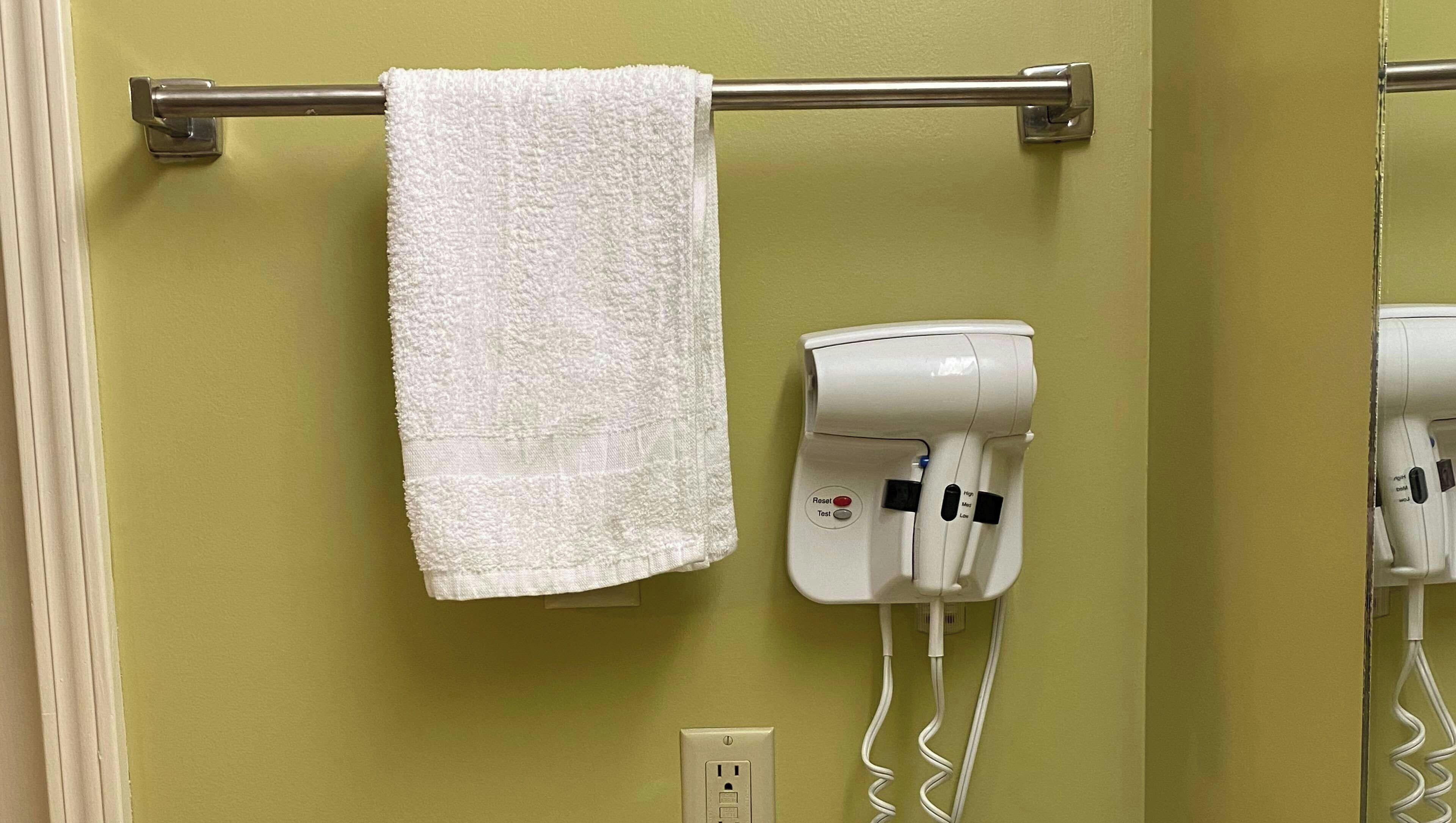 A bathroom with a towel hanging on a metal rod, a hairdryer mounted on the wall, and an electrical outlet below.