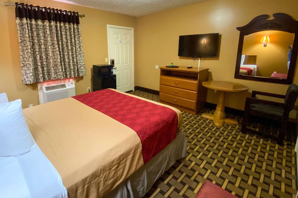 A hotel room with a bed, a desk, a chair, a television, a mirror, and a patterned floor.