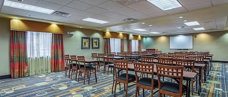 A conference room with rows of wooden chairs and tables, a patterned carpet, and a projector screen.