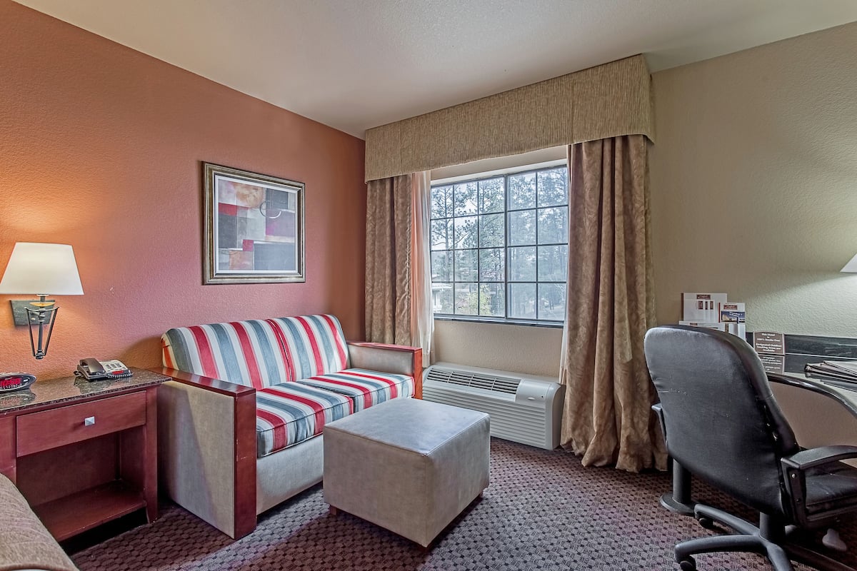 Une chambre d’hôtel avec un canapé à rayures, un petit pouf, un bureau avec une lampe, une chaise et une fenêtre avec des rideaux.