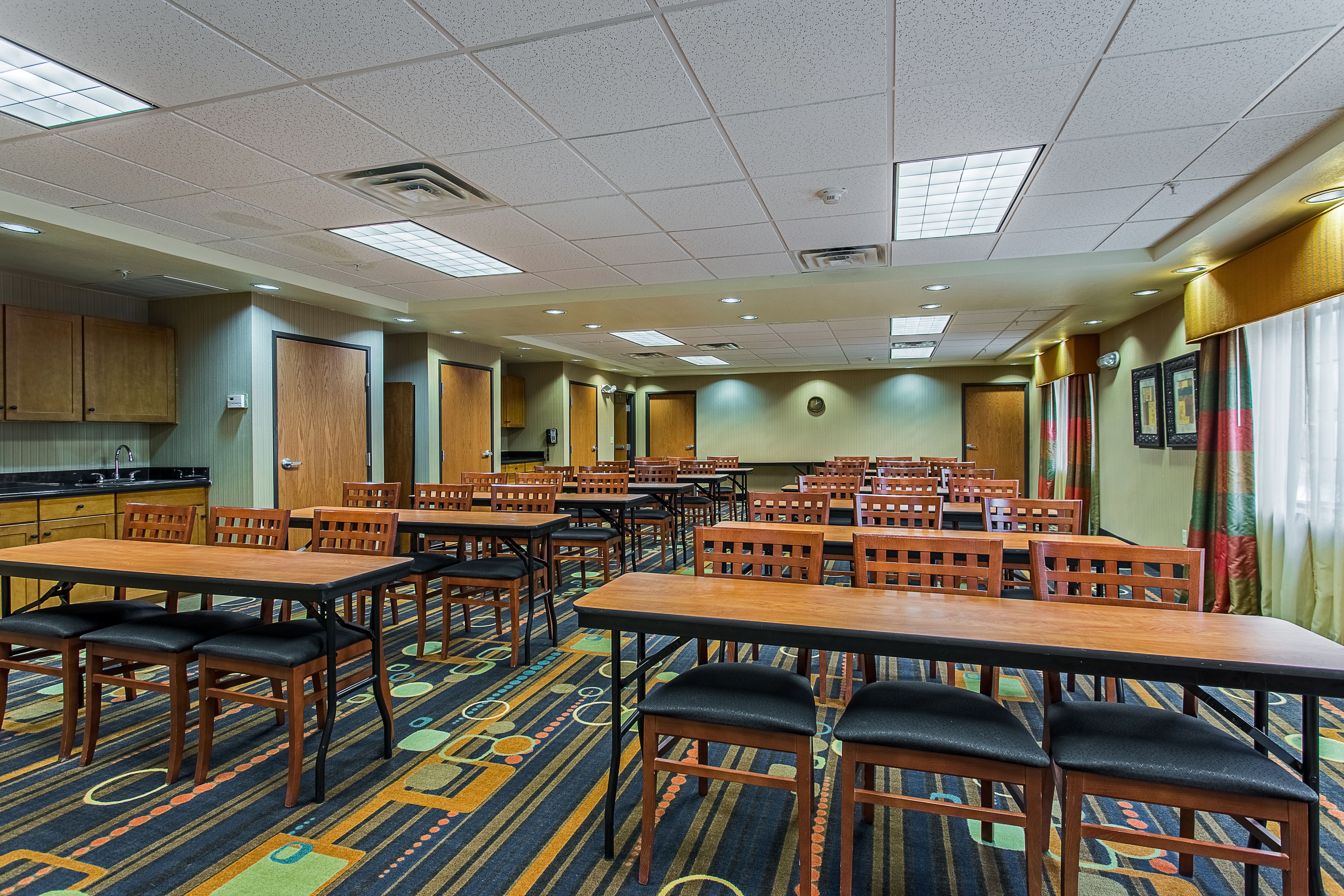 A hotel room with multiple wooden tables and chairs, a patterned carpet, and a kitchenette area.