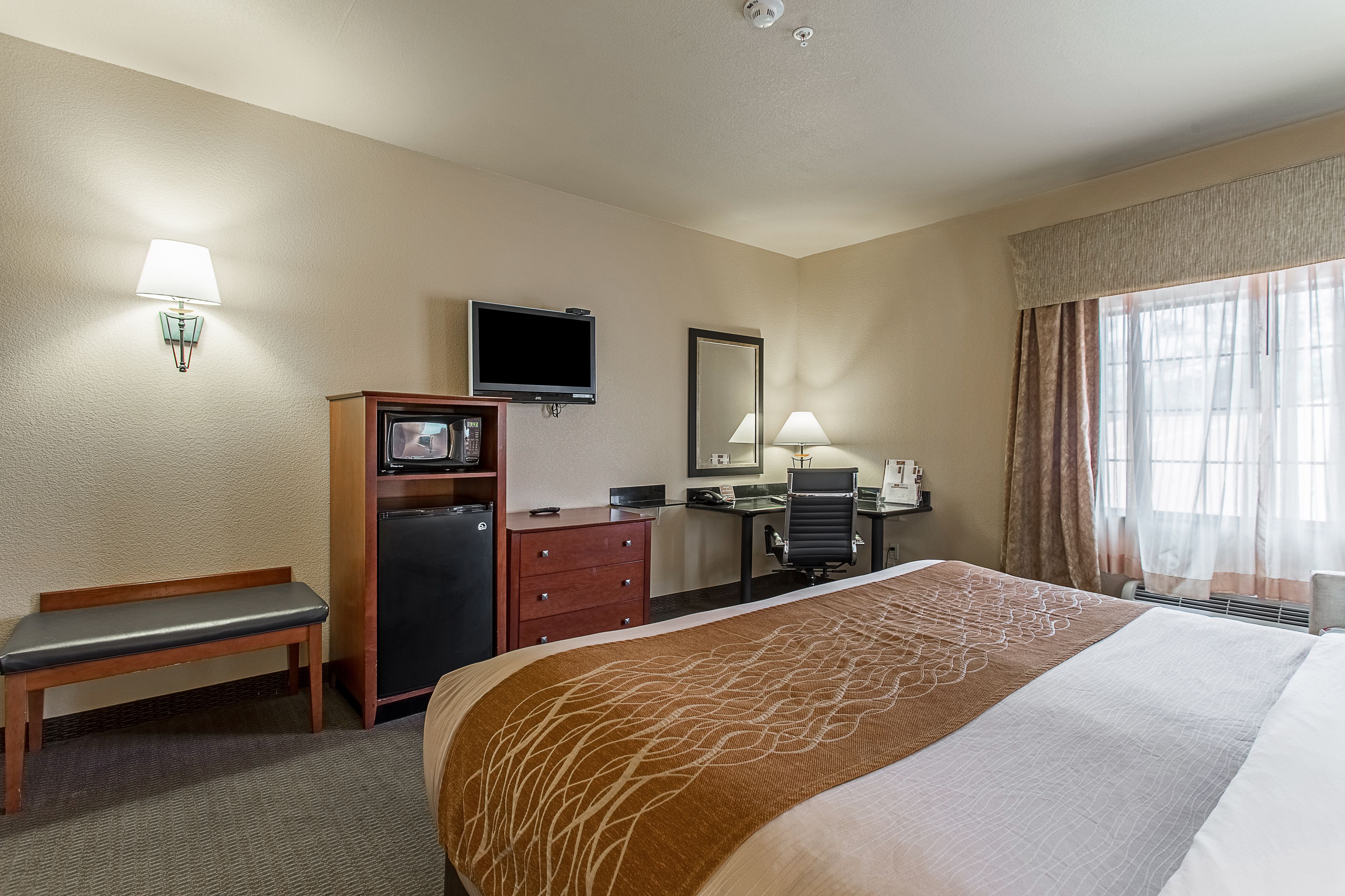 Une chambre d’hôtel avec un grand lit, un bureau avec un ordinateur, une télévision, un banc, un petit réfrigérateur et une fenêtre avec des rideaux.
