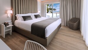 Ein modernes Hotelzimmer mit einem großen Bett, einem Nachttisch mit Lampe, einem Sessel und Blick auf den Strand durch das Fenster.