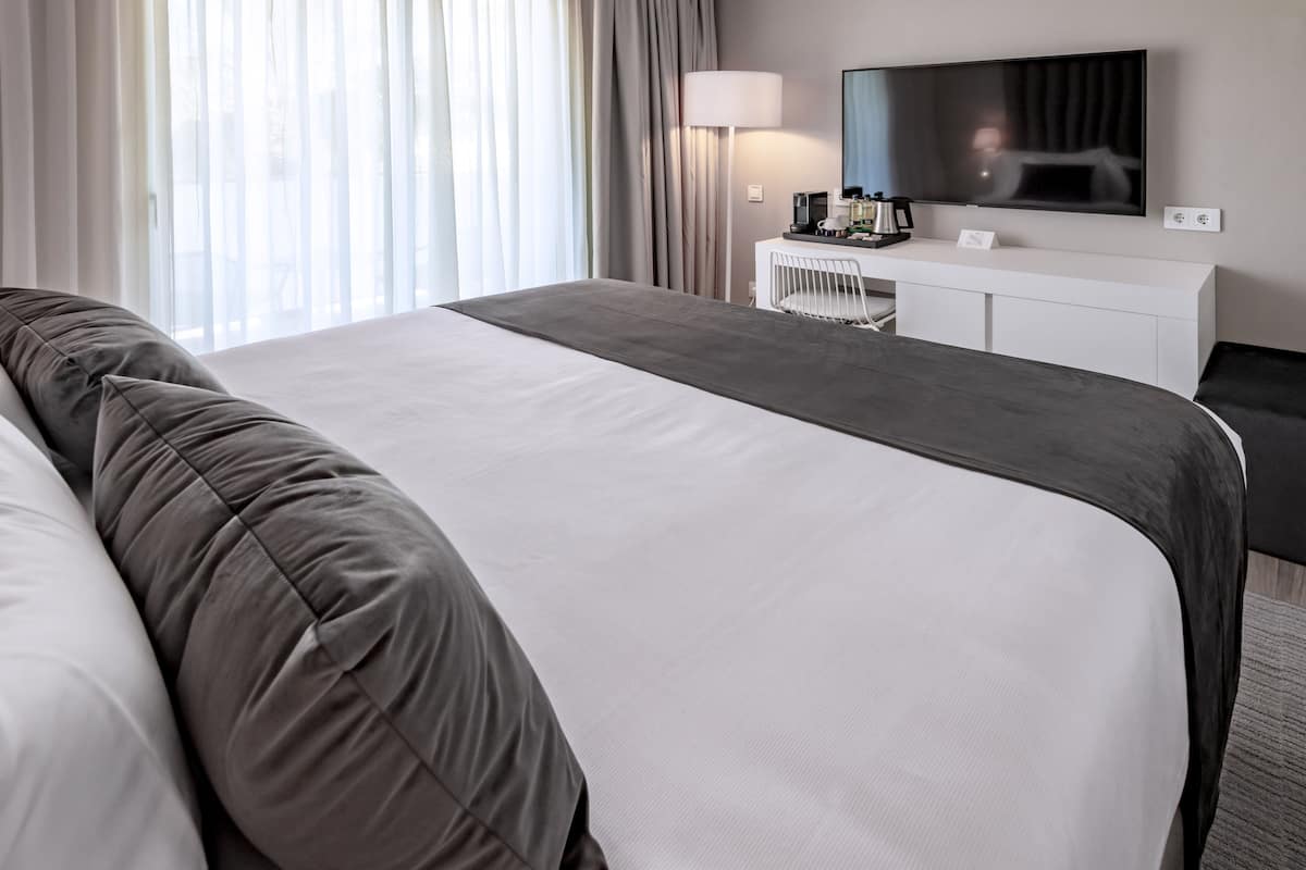 A modern hotel room with a large bed, a bedside table, a flat-screen TV, and a desk with a lamp.