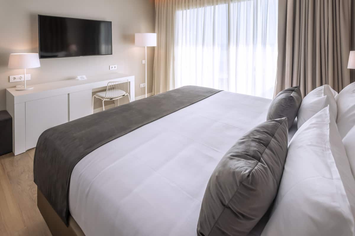 A modern hotel room with a large bed, a flat-screen TV mounted on the wall, a desk with a chair, and a large window with curtains.
