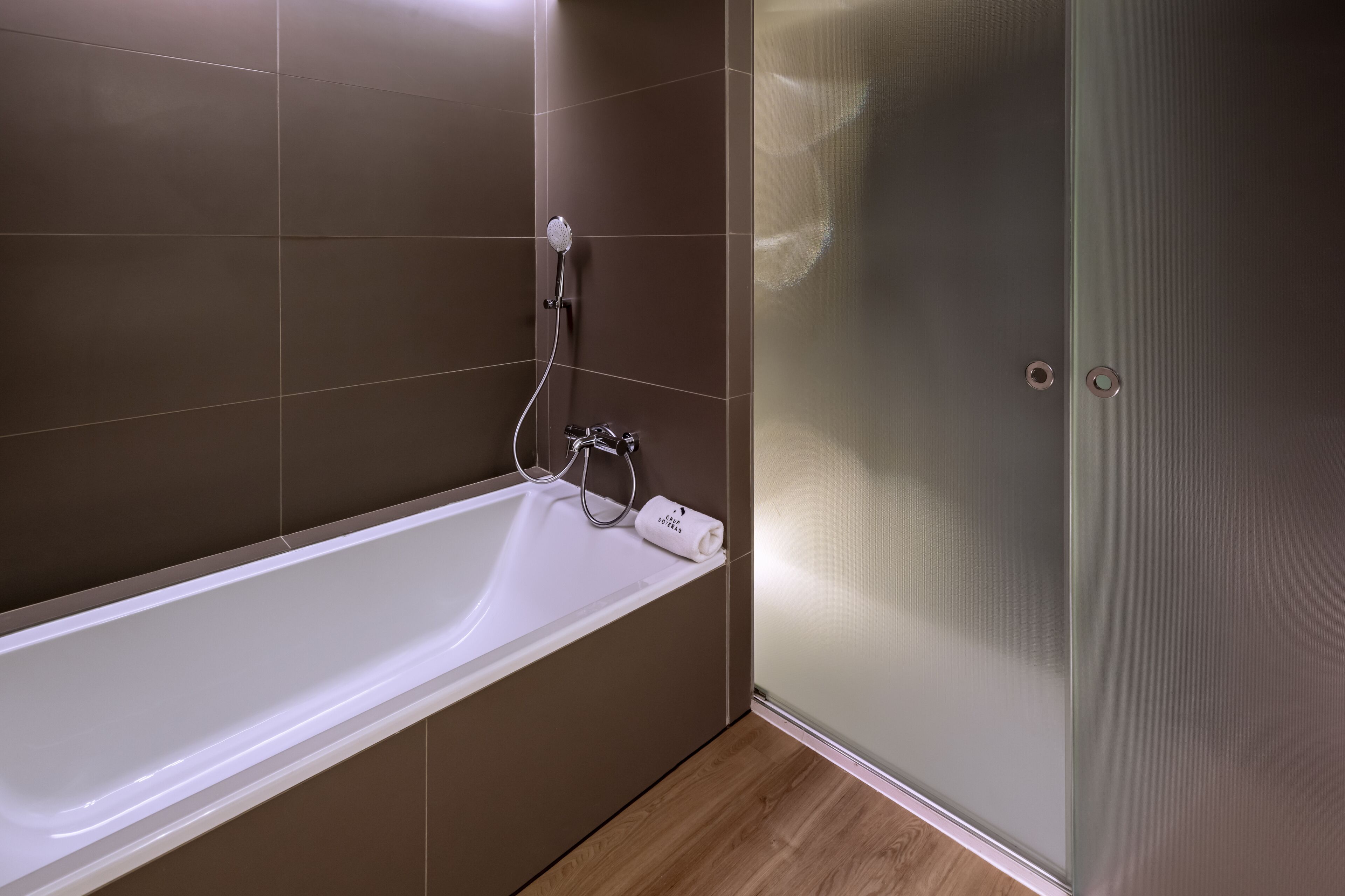 Ein modernes Badezimmer mit einer weißen Badewanne, einer Duschkabine aus Glas und braunen Fliesen.