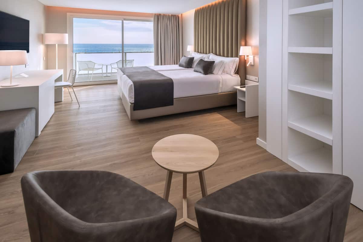 A modern hotel room with a large bed, a desk, two armchairs, and a balcony with a view of the sea.