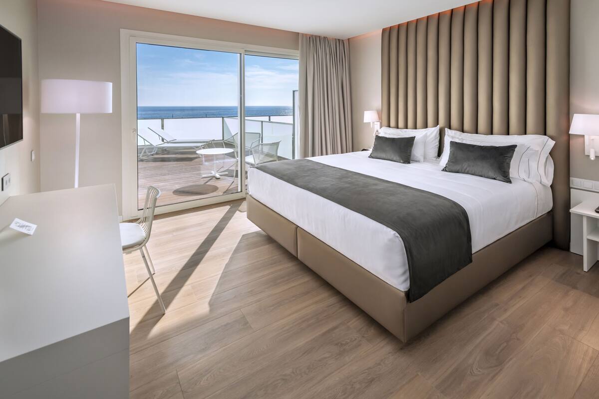 A modern hotel room with a large bed, a balcony with a view of the sea, and a desk with a chair.