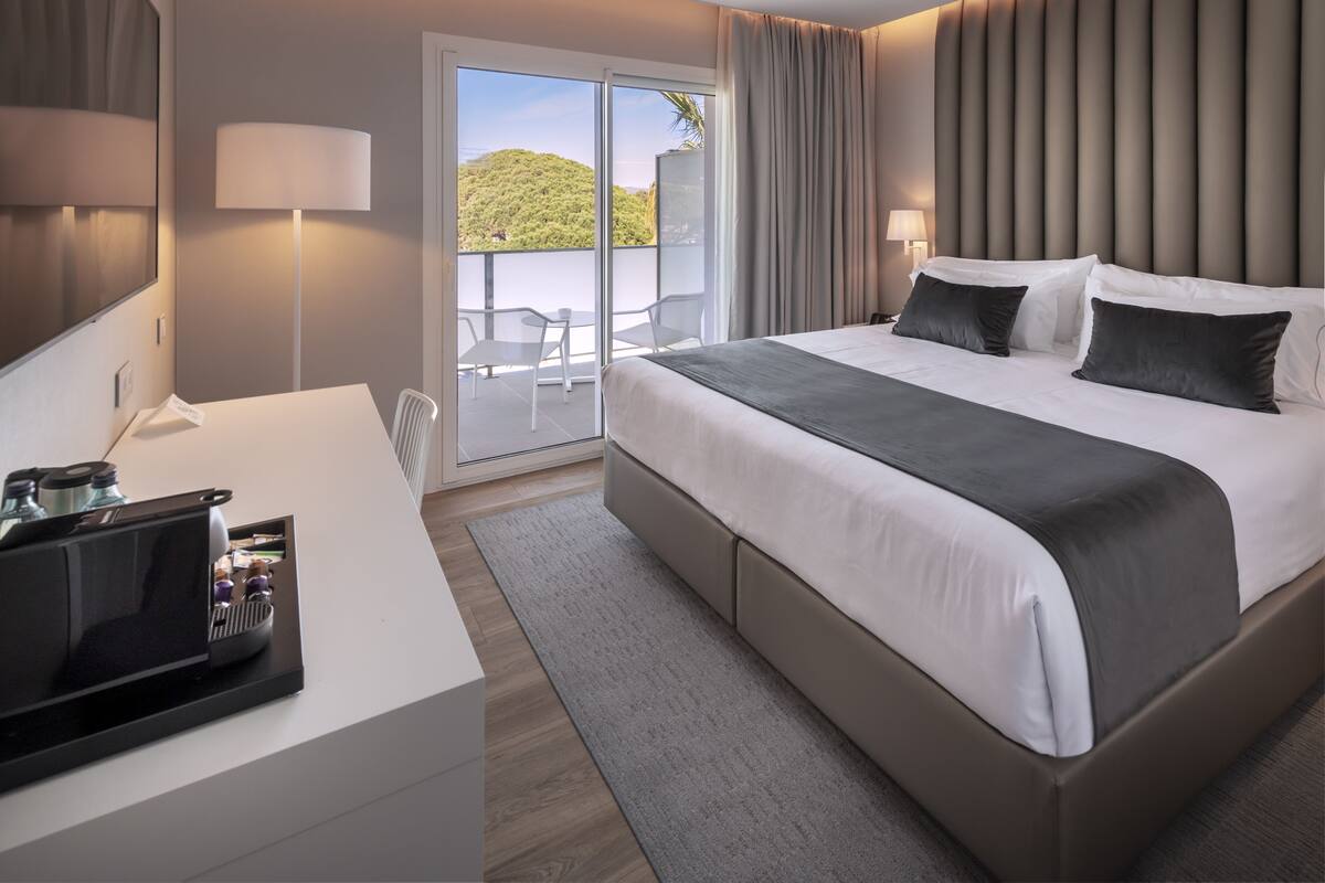 A modern hotel room with a large bed, a desk with a laptop, and a balcony with a view of greenery.