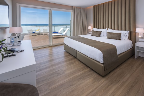Una habitación de hotel moderna con una cama grande, un escritorio con lámpara y vista a la playa a través de una puerta corrediza.