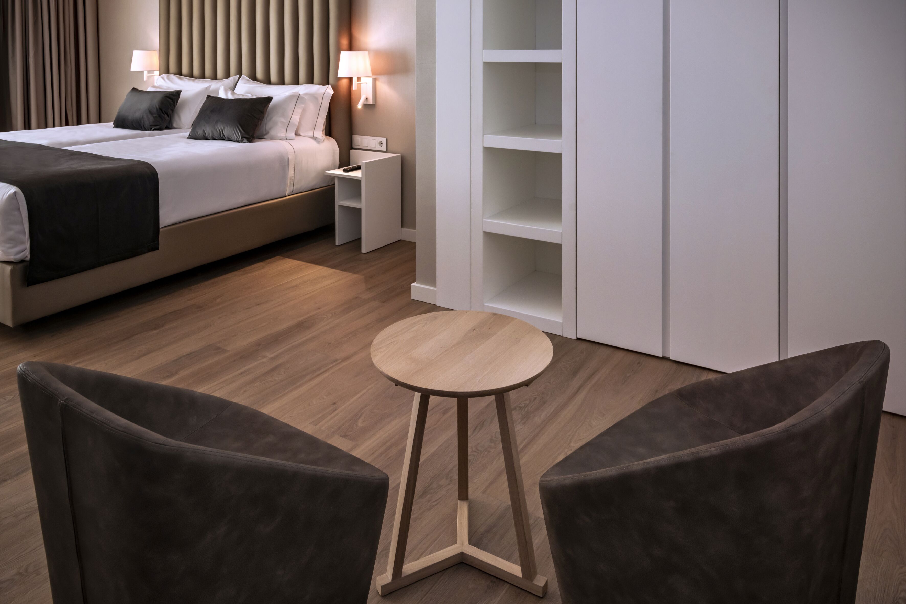 A modern hotel room with a bed, bedside tables, a round wooden table, and two armchairs.