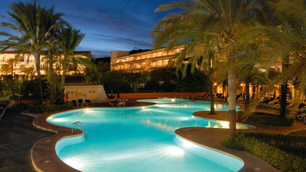 A swimming pool with a curved design, surrounded by palm trees and a building with multiple balconies. - FERGUS Club Font de Sa Cala Beach (Capdepera)