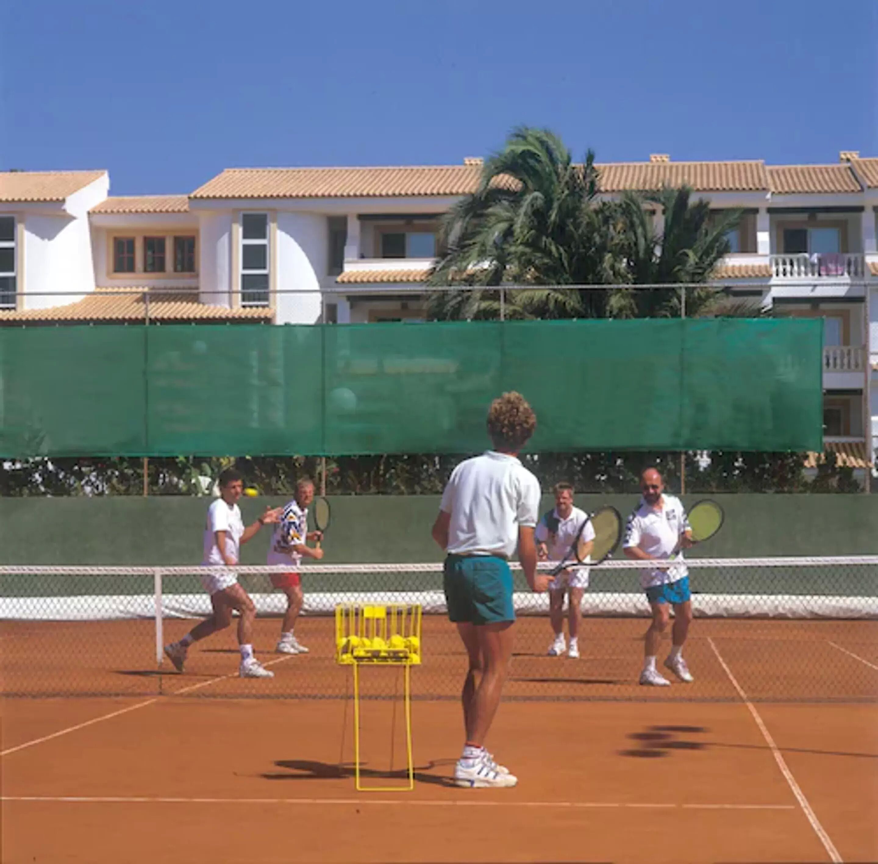 En tennisbane med spillere, et net og et grønt hegn.