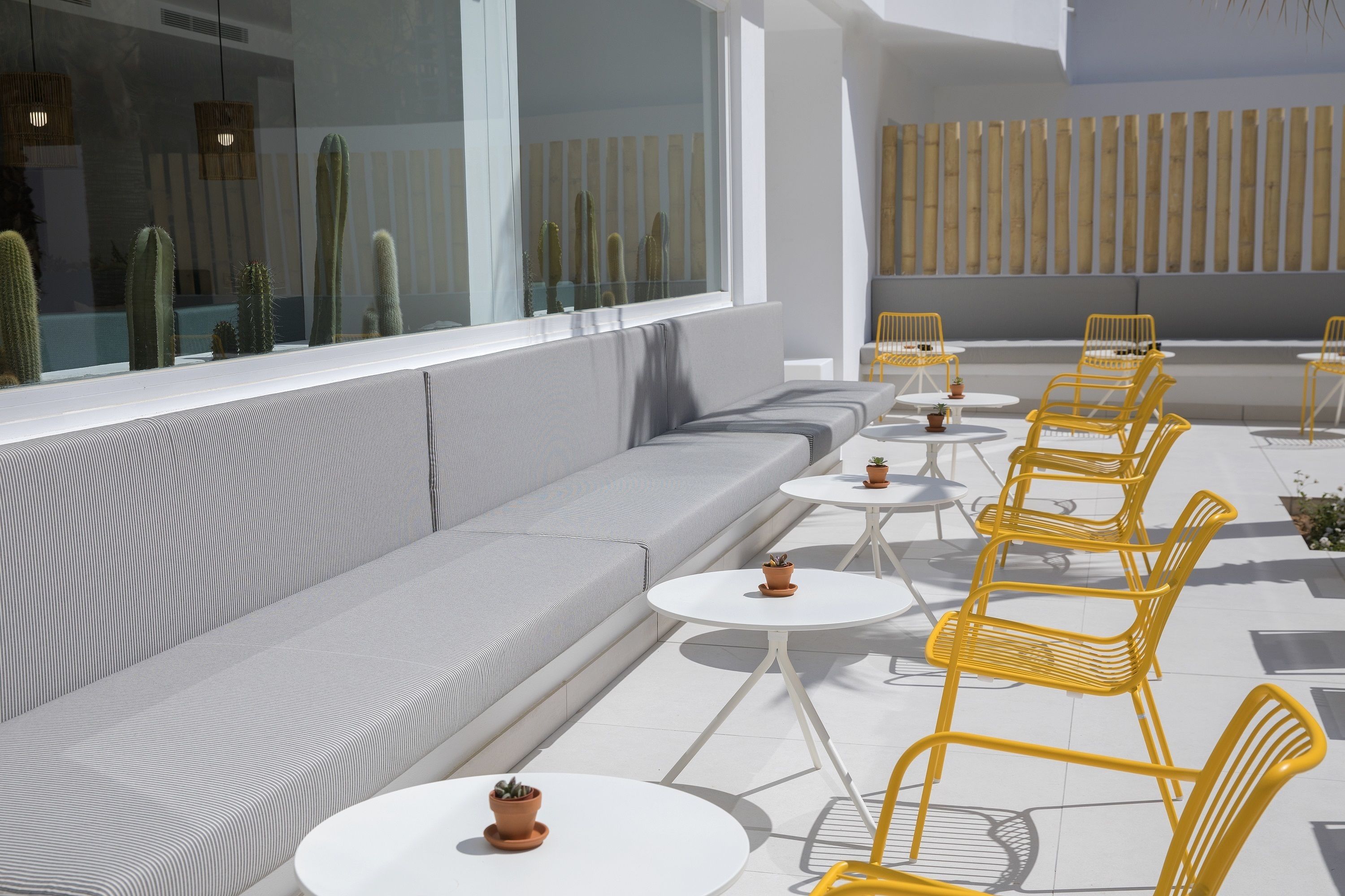 A modern outdoor seating area with a sofa and yellow chairs, white tables, and potted plants.