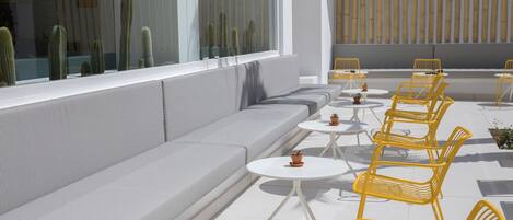 A modern outdoor seating area with a sofa and yellow chairs, white tables, and potted plants.