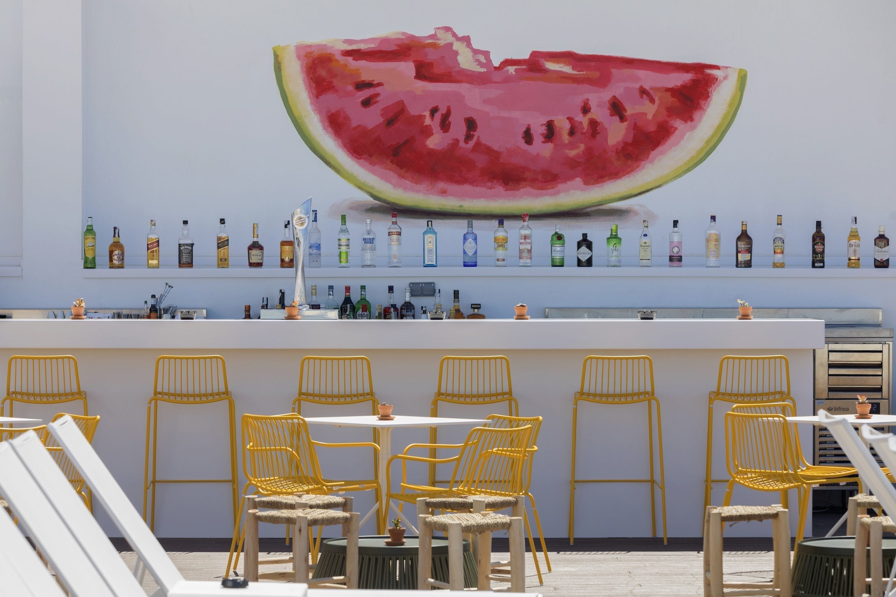 A bar area with a large watermelon mural, yellow chairs, and a variety of liquor bottles on shelves.