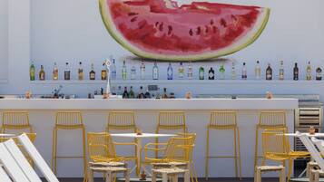 A bar area with a large watermelon mural, yellow chairs, and a variety of liquor bottles on shelves.