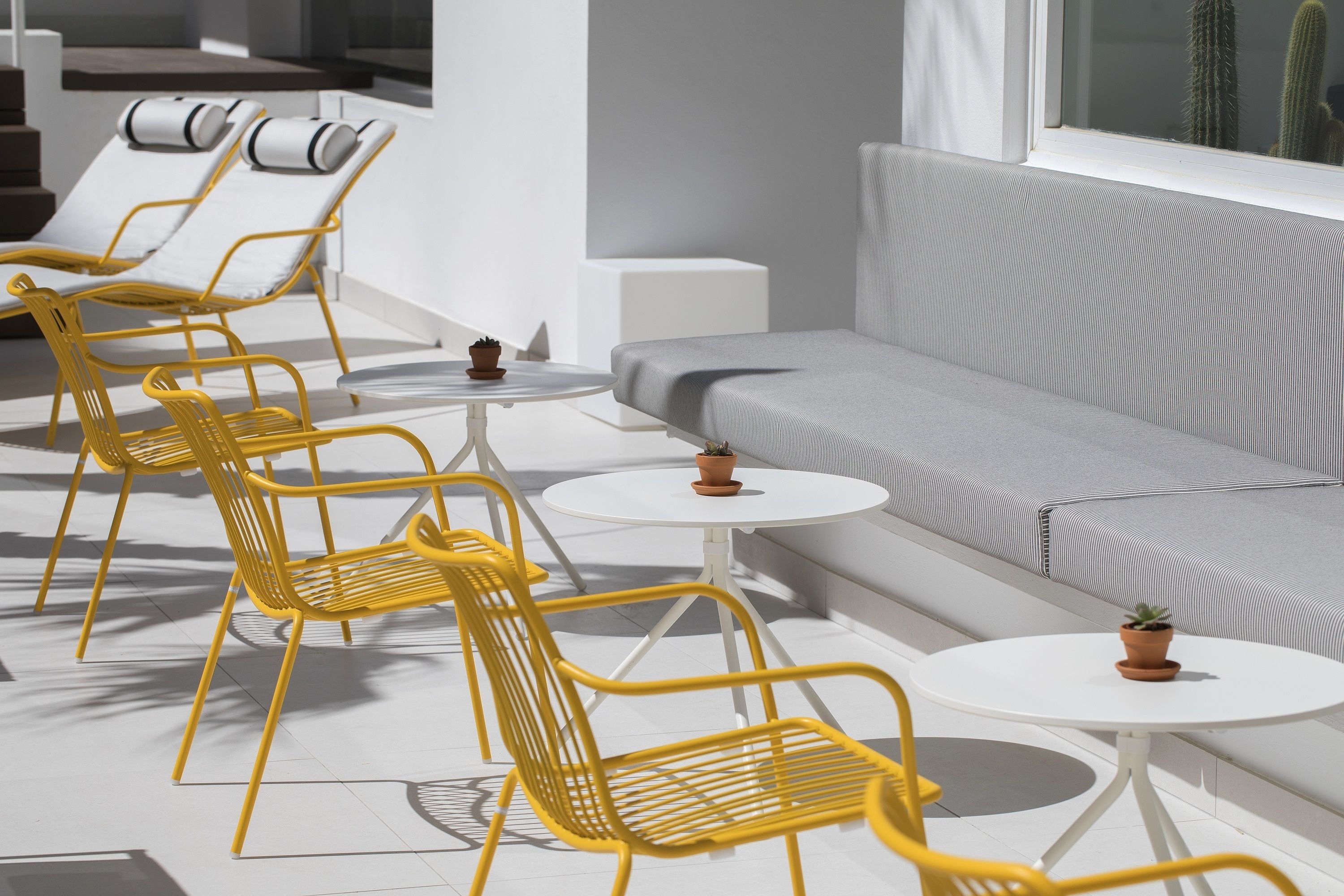Un espace extérieur moderne, aménagé avec des chaises jaunes et des tables blanches, un canapé gris et un cactus en arrière-plan.
