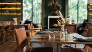 A restaurant interior with wooden tables and chairs, a vase with dried flowers, and a fireplace in the background. - Paperbark Camp (Woollamia)