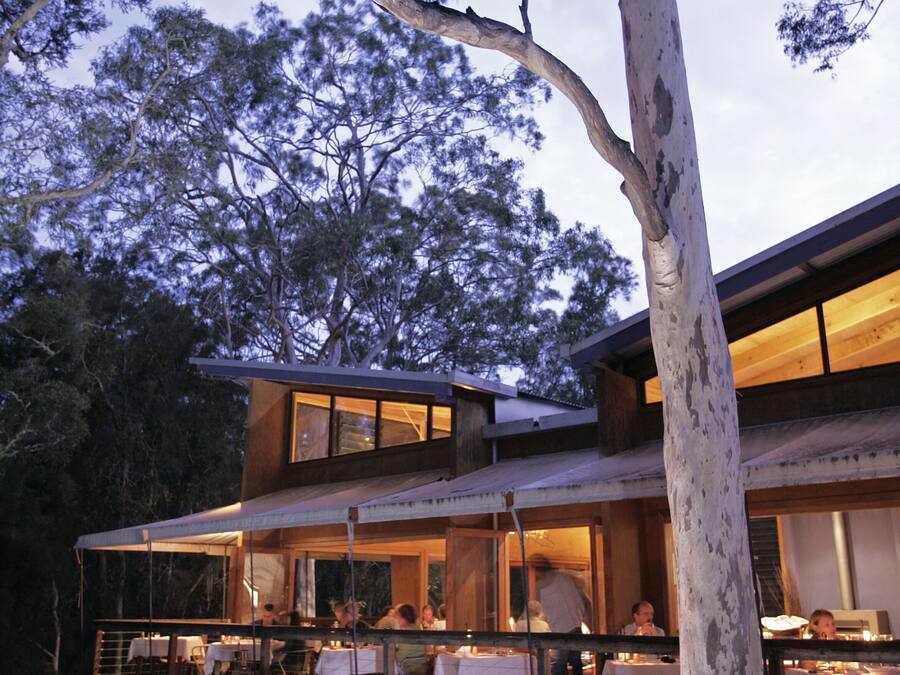 A modern building with a dining area on an elevated deck, surrounded by trees.