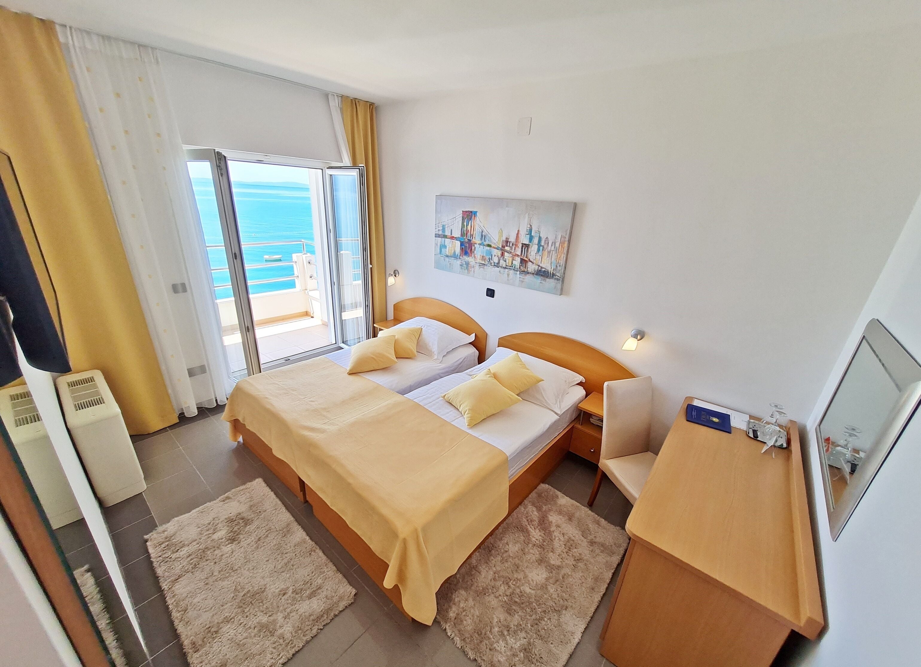 A hotel room with a bed, bedside tables, a desk, a chair, a painting, and a view of the sea through a sliding door.
