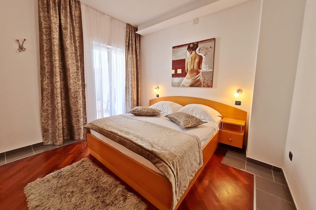 A bedroom with a wooden bed, patterned curtains, a painting on the wall, and a nightstand with a lamp.