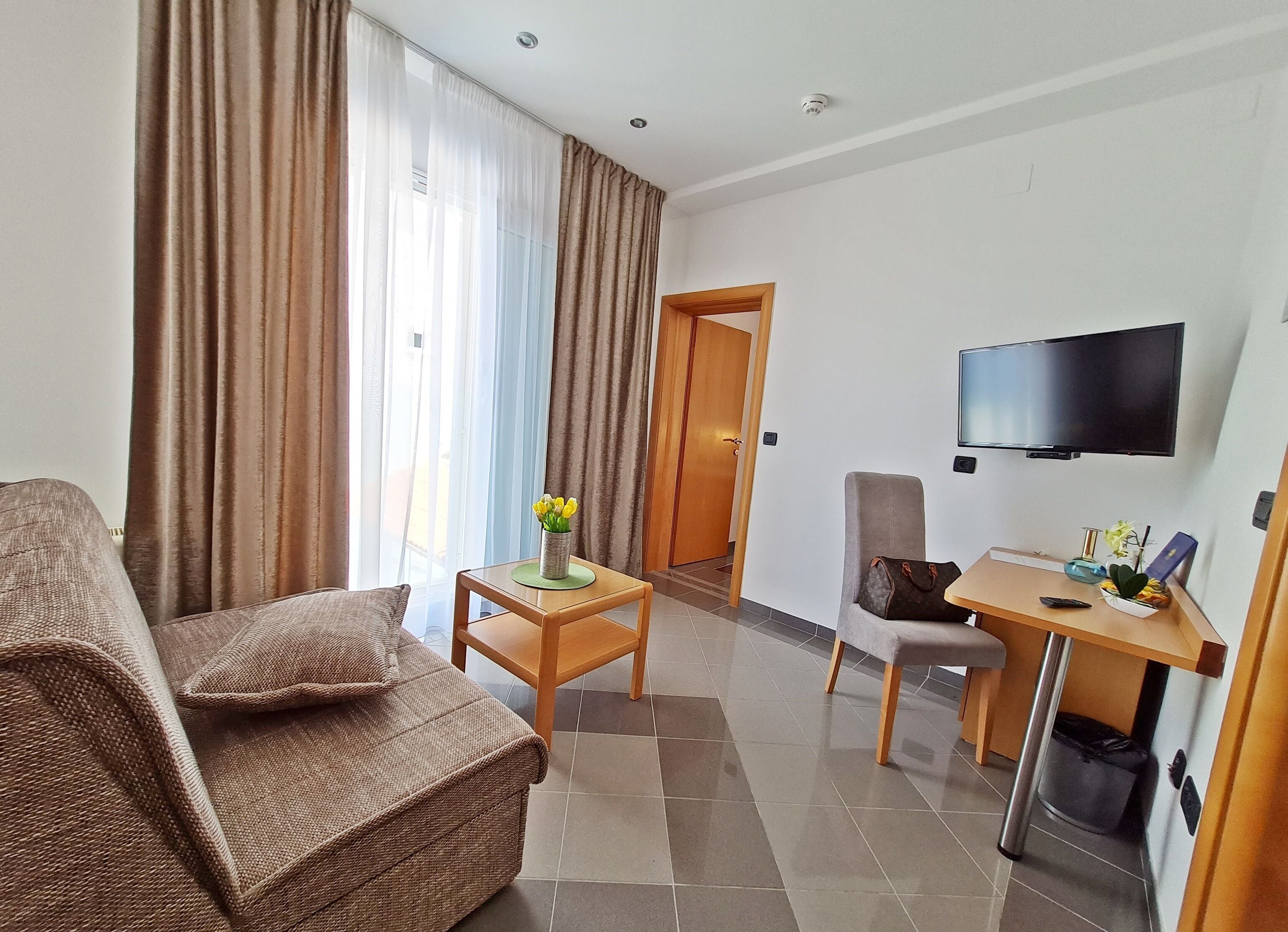 Une chambre d’hôtel avec un canapé, une petite table ornée d’un vase de fleurs, un bureau avec une chaise, un téléviseur fixé au mur et une porte donnant sur une autre pièce.