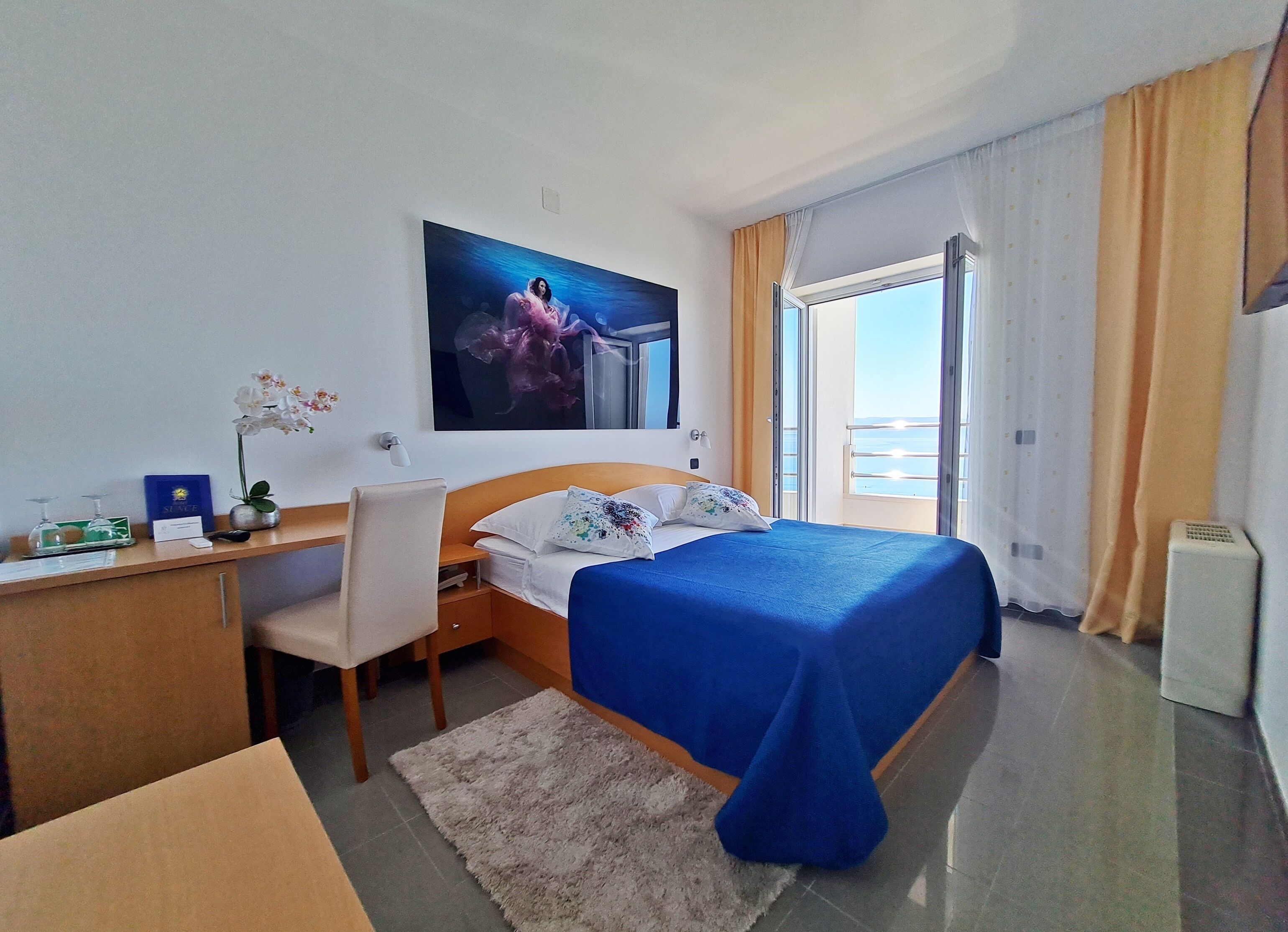 Habitación de hotel con cama, escritorio, silla y un amplio ventanal con vista al mar.