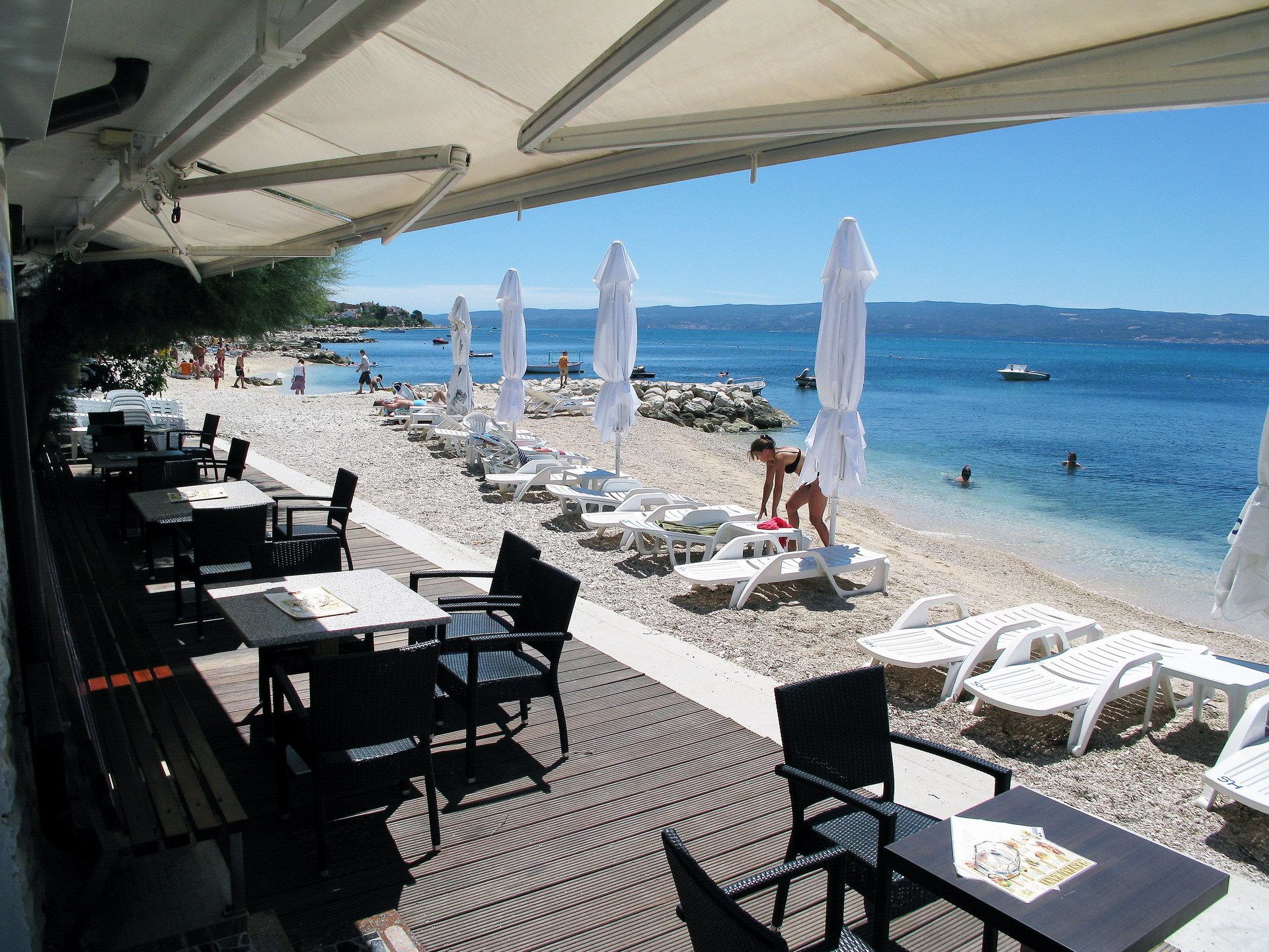 A beachside restaurant with outdoor seating, white umbrellas, and a view of the beach and sea.