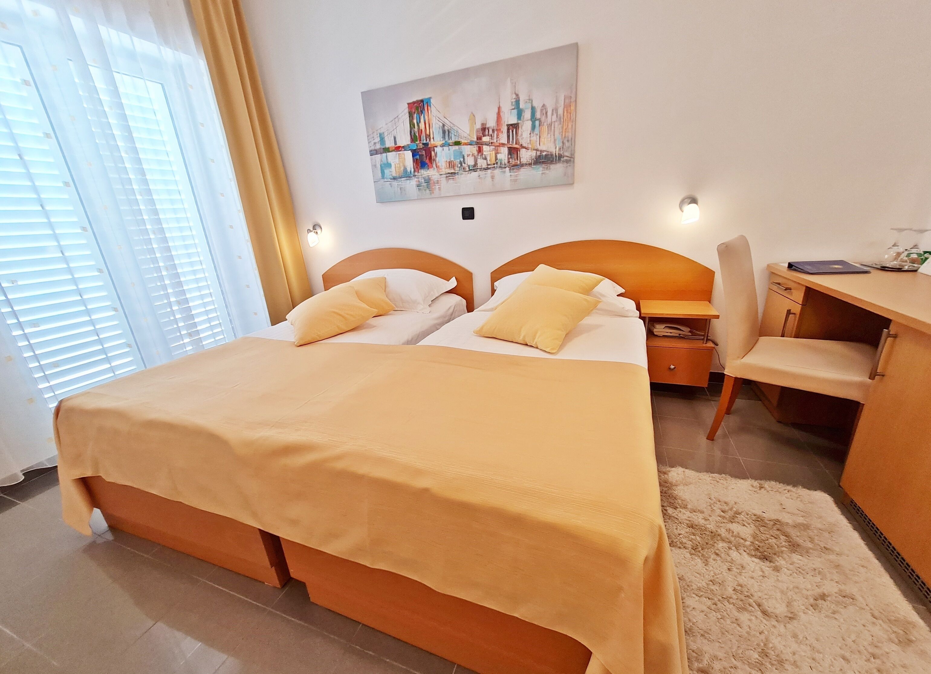 Ein Hotelzimmer mit Bett, Schreibtisch und Stuhl. An der Wand hängt ein Bild, und ein Fenster mit Jalousien.