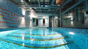 Una piscina interior con mosaicos azules, techo de cristal y una escalera que lleva a una puerta.