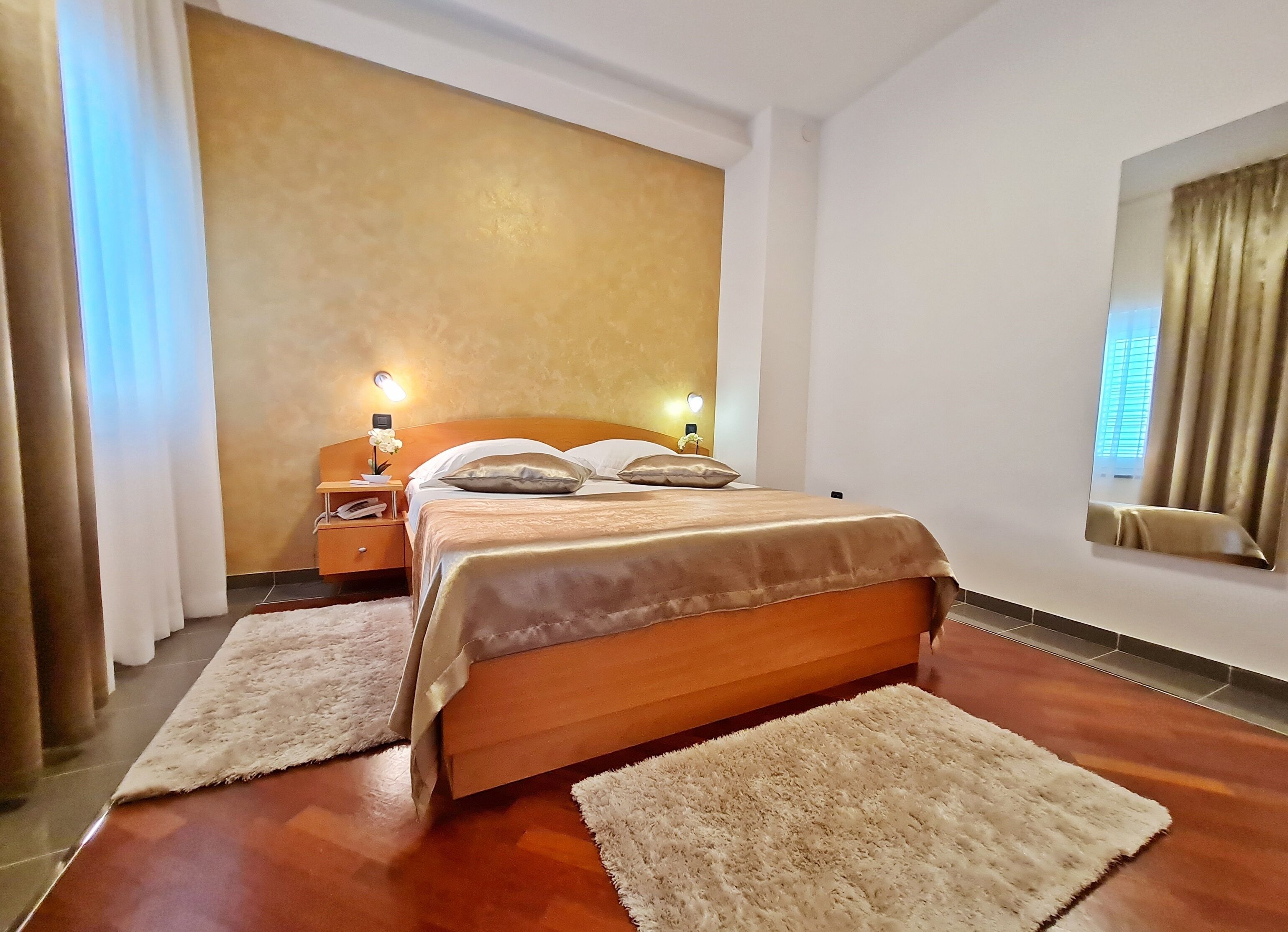 A hotel room with a large bed, two bedside tables, a nightstand, a wall-mounted mirror, and a window with curtains.