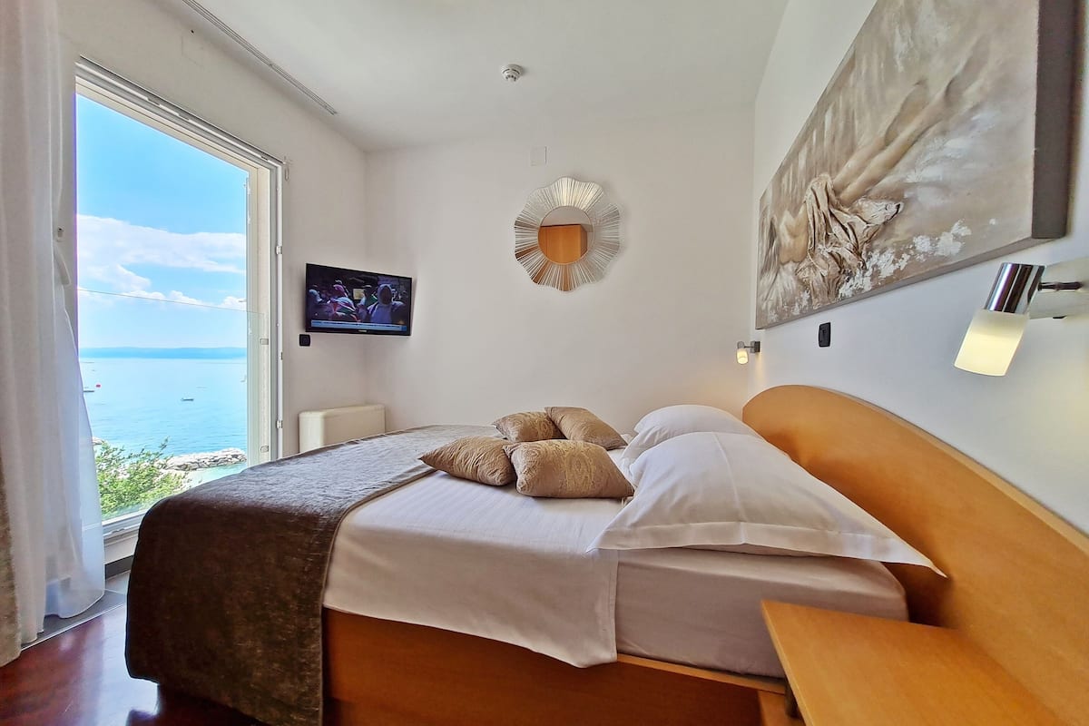 A bedroom with a bed, a TV, a painting, and a view of the sea.