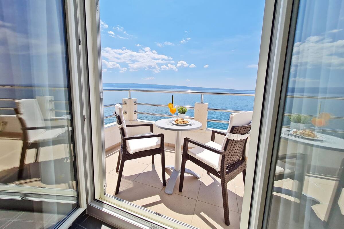 A balcony with wicker chairs and a small table, overlooking a body of water and distant land.