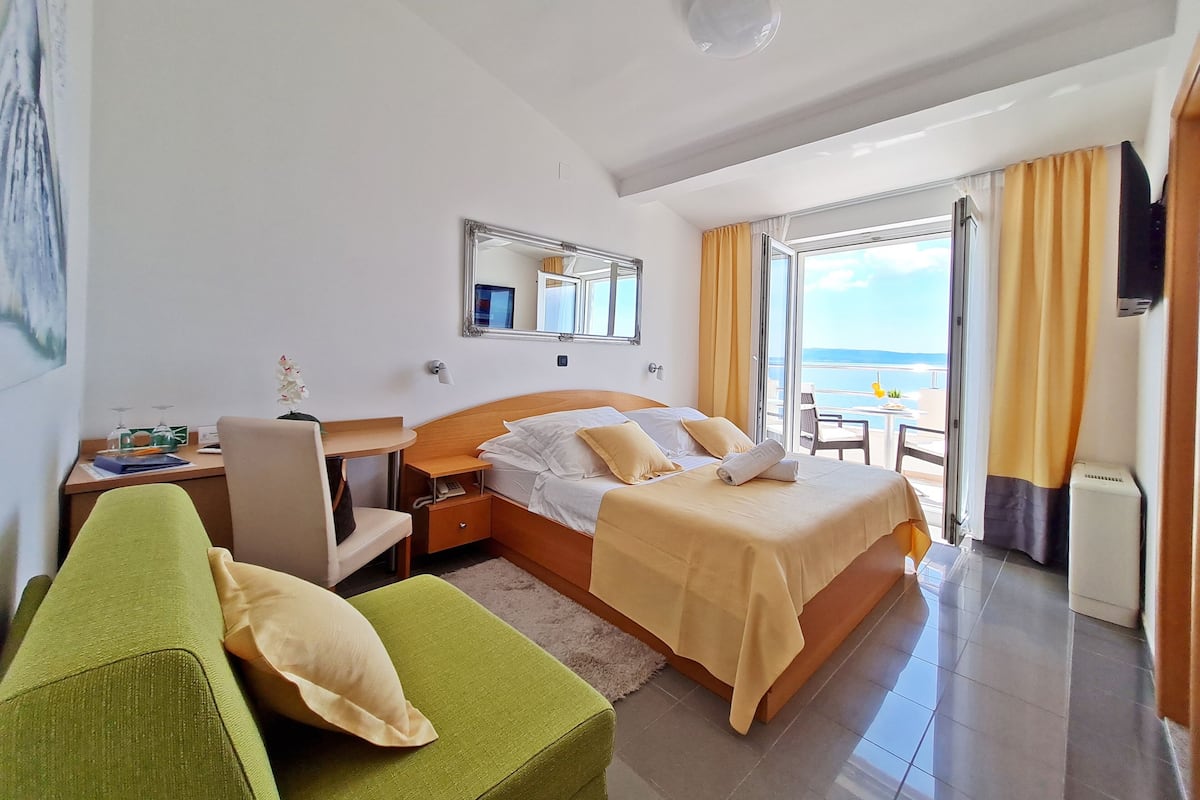 A hotel room with a bed, a desk, a chair, a sofa, and a balcony with a view of the sea.