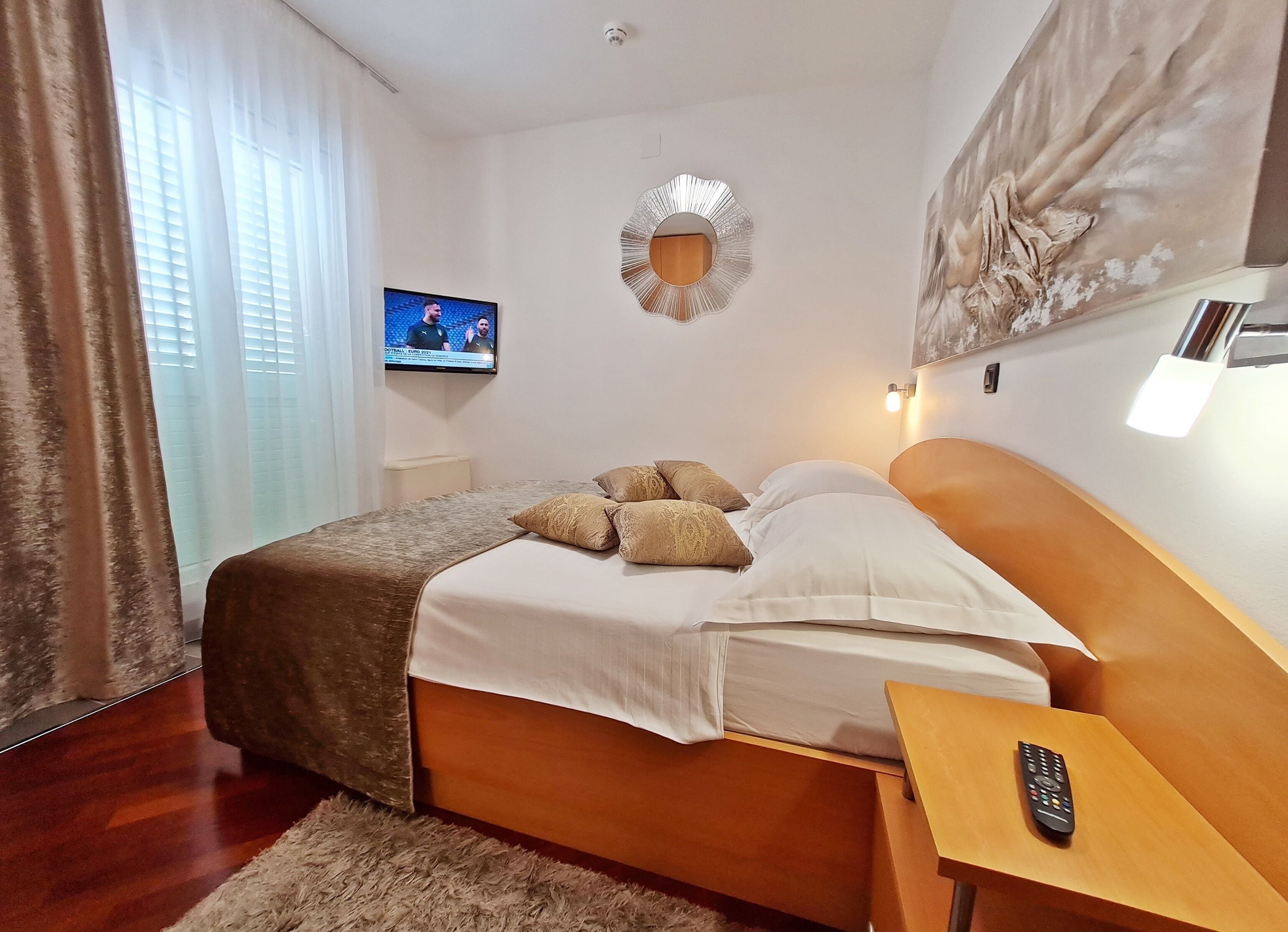 Habitación de hotel con cama, televisor montado en la pared, un cuadro y control remoto sobre una mesita de noche.