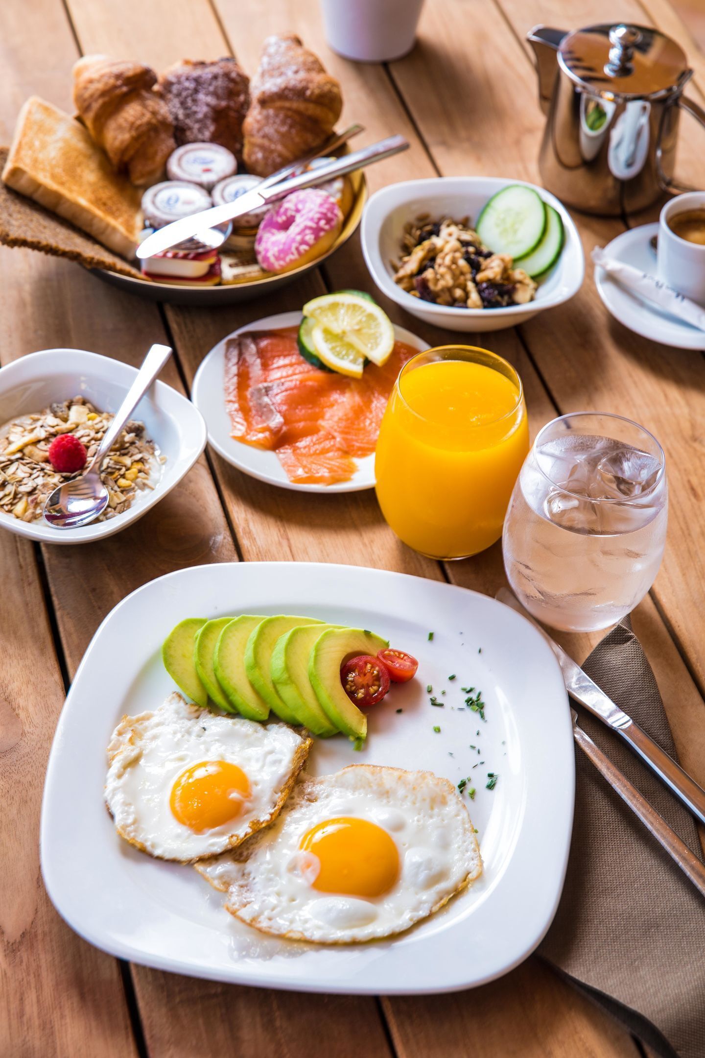 A breakfast setting with eggs, avocado, toast, croissants, and beverages on a wooden table.