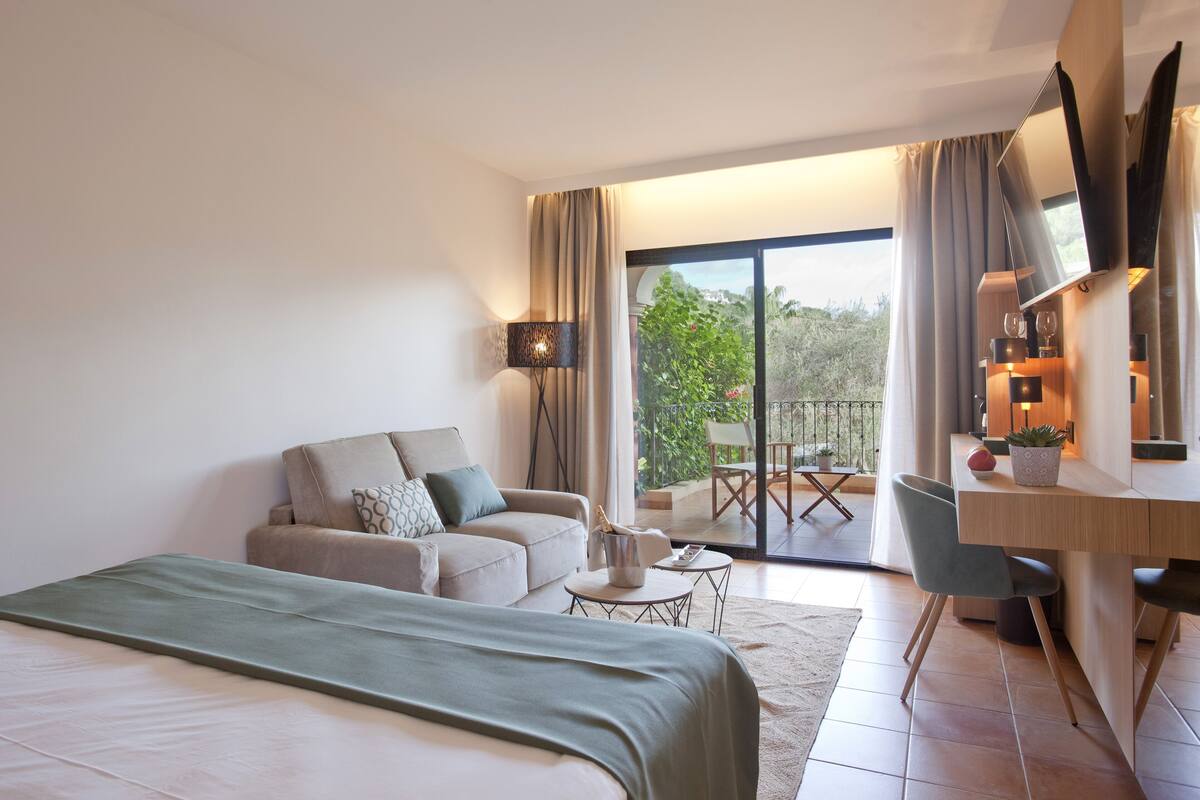 A modern hotel room with a bed, a sofa, a desk, and a balcony with a view of greenery.