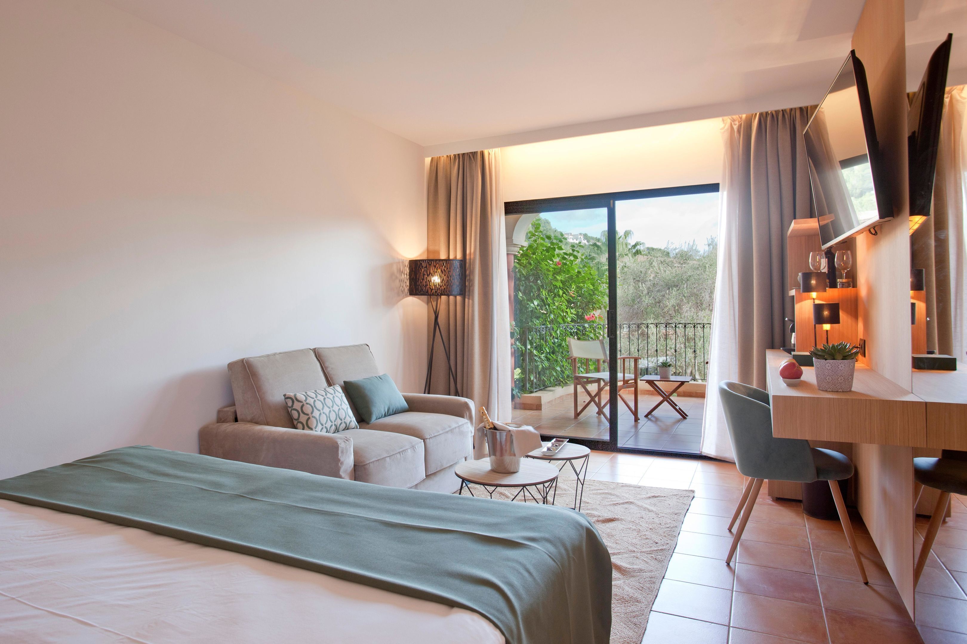A modern hotel room with a bed, a sofa, a desk, a chair, a TV, and a balcony with a view.