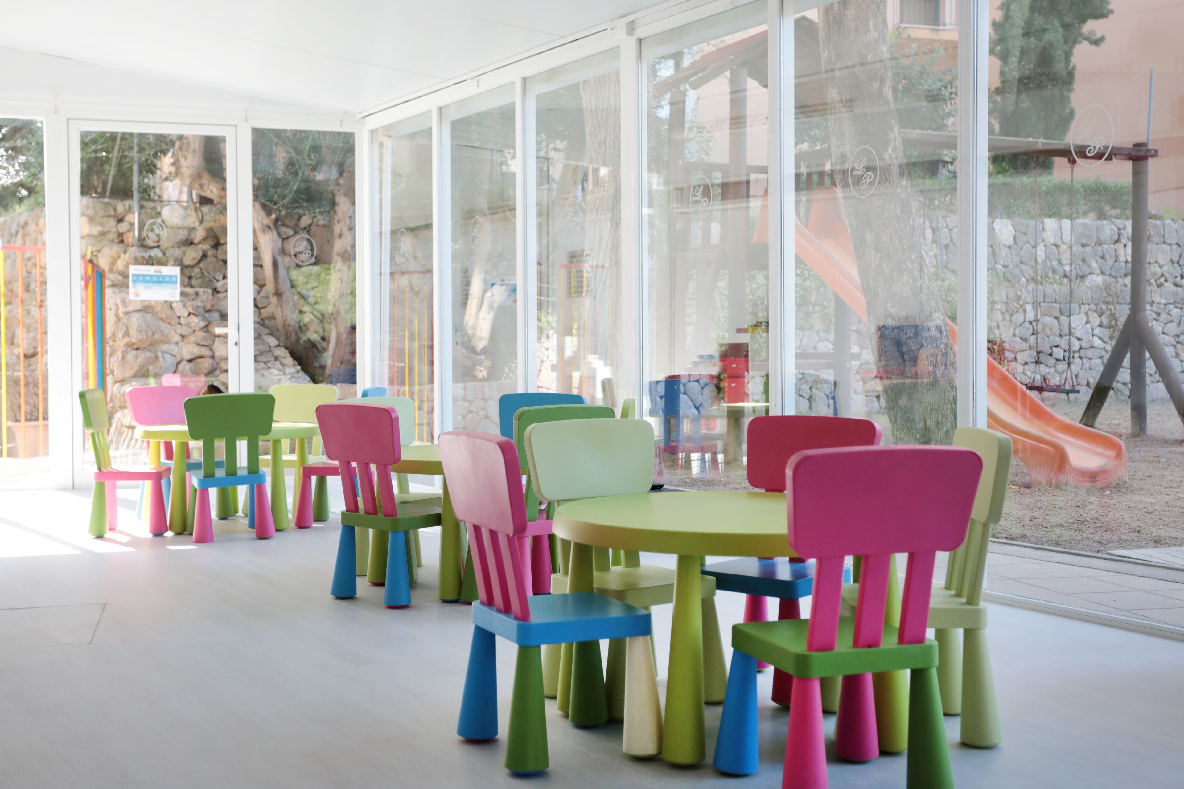 A children's play area with colorful chairs and tables, large windows, and a view of a playground outside.