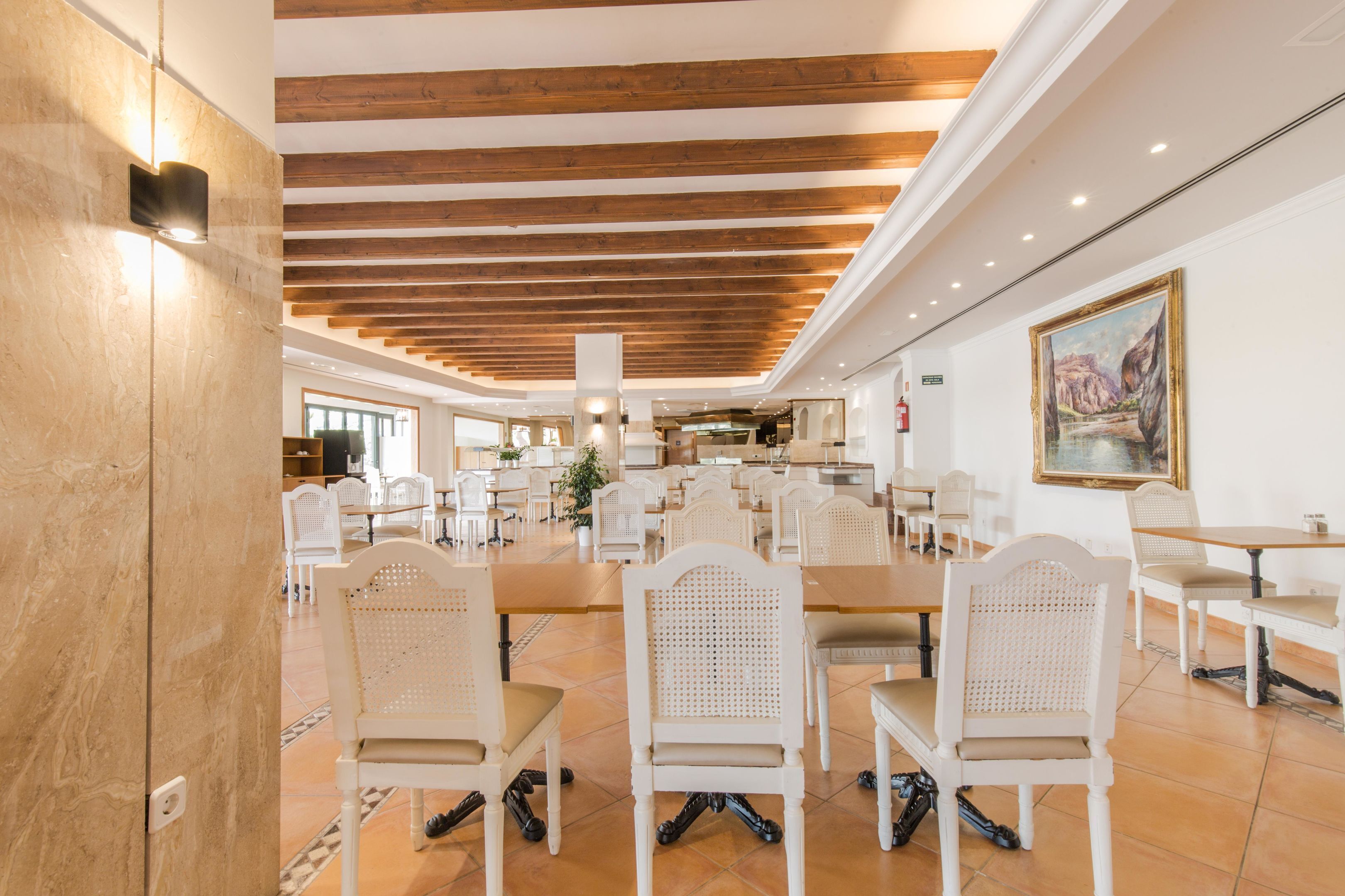 A spacious dining area with white chairs, a long table, wooden beams on the ceiling, and a painting on the wall.