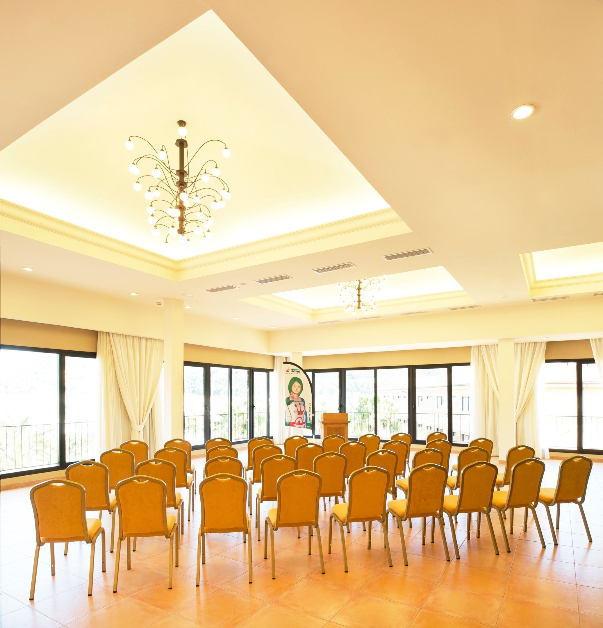 A conference room with rows of chairs, a chandelier, large windows, and a podium.