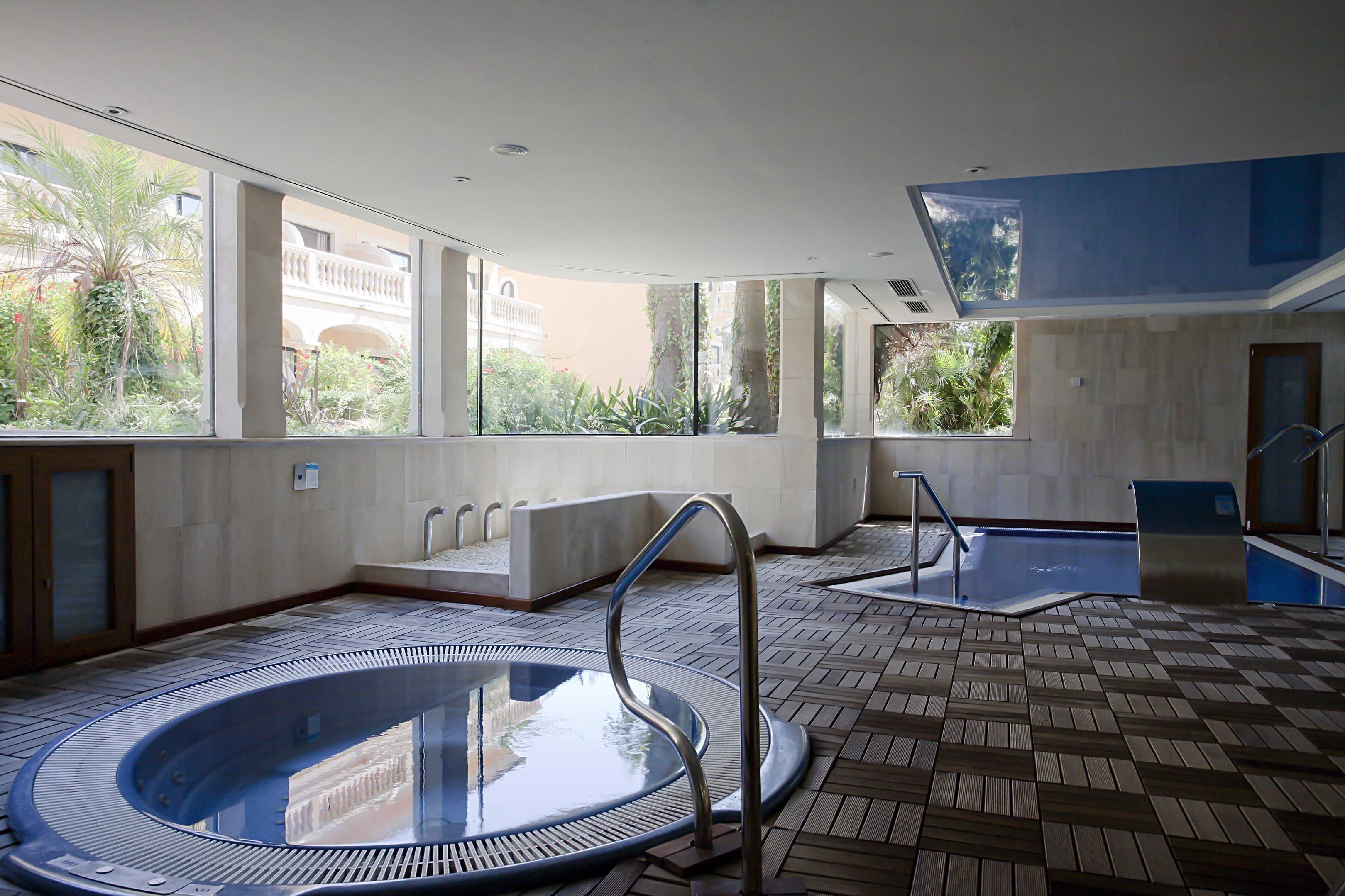 Un espace piscine intérieur moderne, doté d’un jacuzzi à moitié vitré, d’une terrasse ensoleillée offrant une vue sur les bâtiments environnants et d’un sol carrelé.