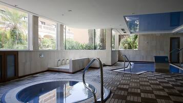 Un espace piscine intérieur moderne, doté d’un jacuzzi à moitié vitré, d’une terrasse ensoleillée offrant une vue sur les bâtiments environnants et d’un sol carrelé.
