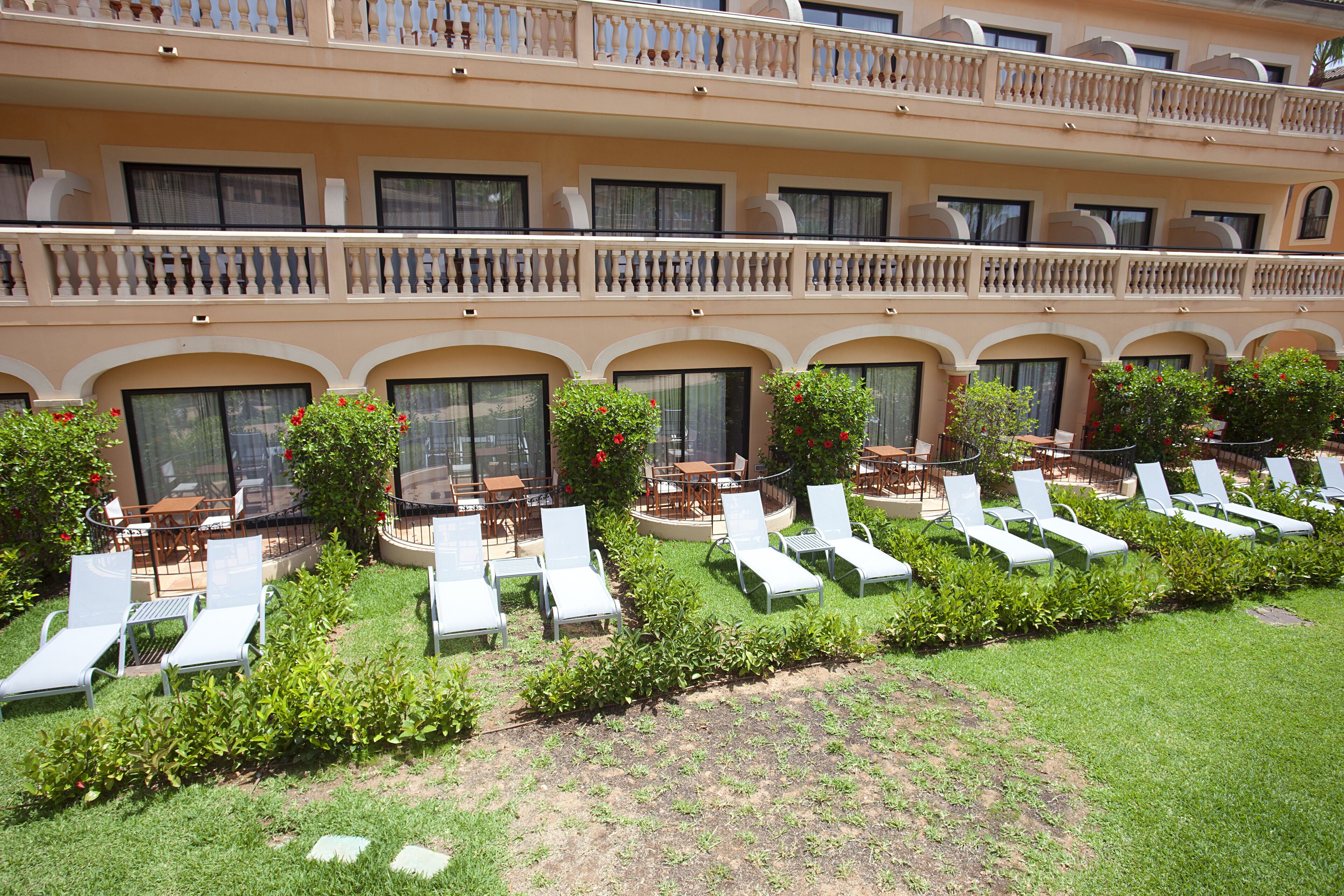 A hotel with multiple balconies, outdoor seating areas with tables and chairs, and a well-maintained lawn with sun loungers.