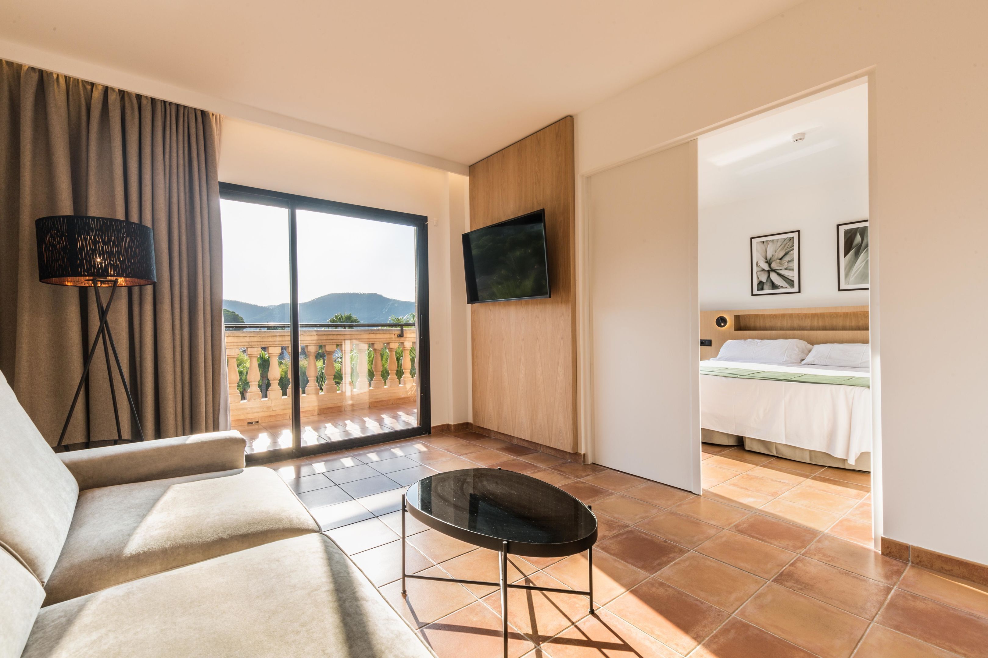 A hotel room with a sofa, a small table, a TV mounted on the wall, and a bed with a view of the mountains.