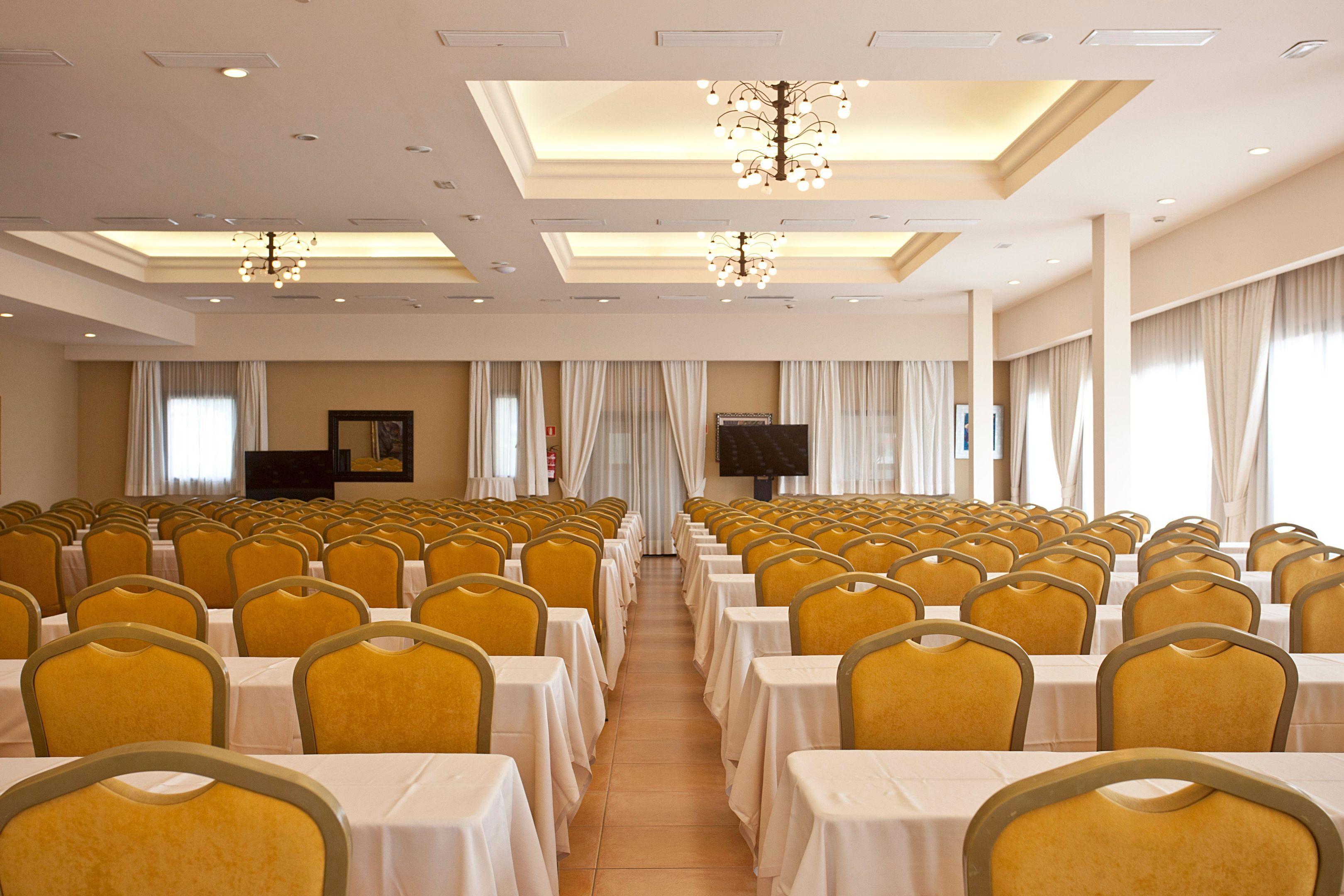 Un salón de conferencias con filas de mesas y sillas, una araña y una pantalla de televisión.