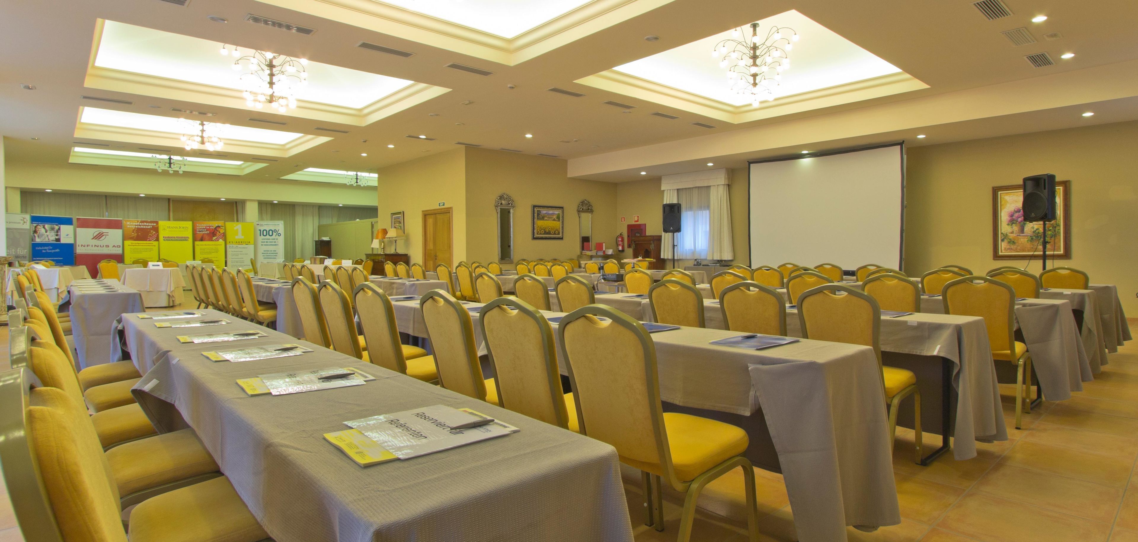 Un salón de conferencias con filas de mesas y sillas, una pantalla para proyector y un altavoz en la pared.