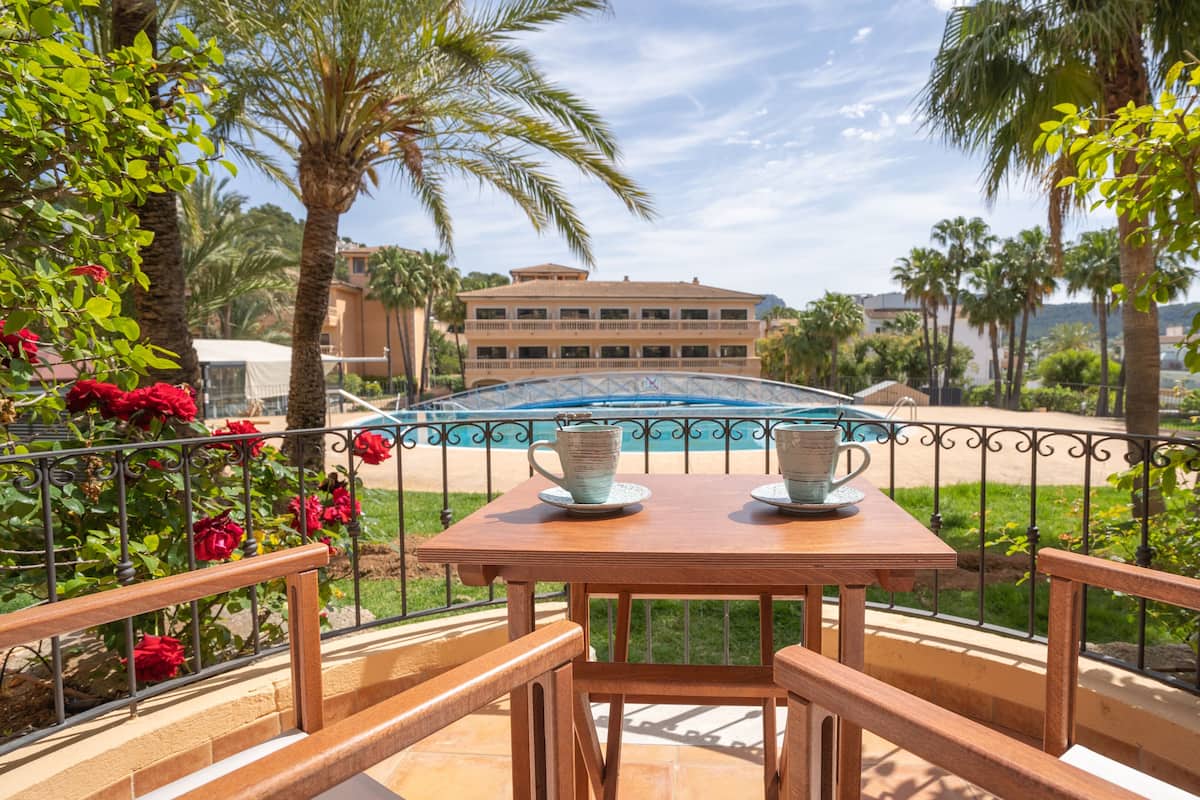 A wooden table with two ceramic cups on a balcony overlooking a swimming pool, surrounded by palm trees and a building in the background.